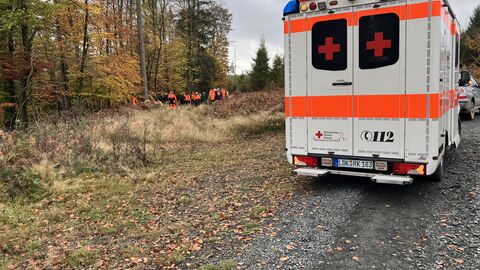 Rettungswagen von hinten auf einem Waldweg. Im Hintergrund erkennt man Menschen am Wald stehend.