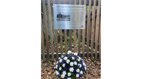Gedenktafel an Zaun befestigt. Davor liegt ein Blumenkranz.