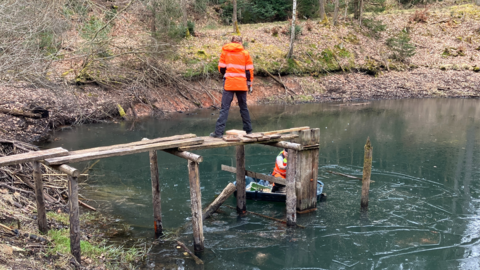Auf dem Foto sehen Sie einen Freiwilligendienstleistenden auf einem Steg. Ein anderer Sitz auf einem Boot auf einem Teich.