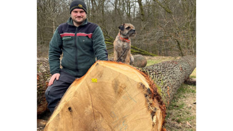 Ein Revierleiter steht mit seinem Hund neben dem teuersten Eichenstamm