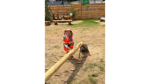 Ein Kind spielt mit Spielzeugmotorsäge, Helm und Warnweste an einem Entastungsspiel.