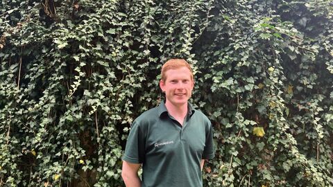 Eine junge männliche Person steht in grünem Dienstpoloshirt vor einer bewachsenen Mauer und blickt den Betrachter an. 