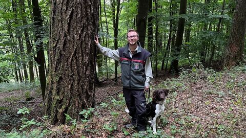 Jan Lennart Fischer mit Hund Ein junger Förster steht mit seinem Jagdhund an einer starken Lärche, berührt sie mit seiner Hand und lächelt in die Kamera.