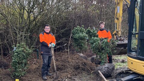 Zwei Personen in Forst-Signalkleidung mit Spaten. Rechts ist ein Bager zu sehen. 