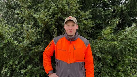 Ein junger Mann mit Signalkleidung blickt den Betrachter an. Er hat den Arm in die Hüfte gebeugt und steht vor jungen Bäumen und büschen. 