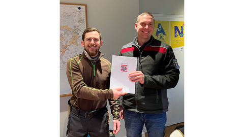 Zwei Männer halten eine Urkunde fest und lachen den Betrachter an. 