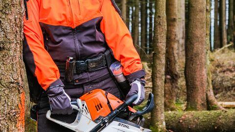 Eine Forstwirtin in Schutzkleidung hält eine Motorsäge und lehnt an einer Fichte. Sie lächelt den Betrachter an. 