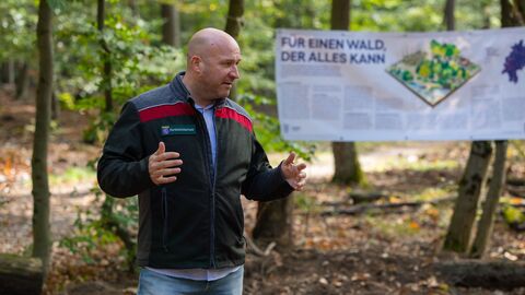 Forstminister Ingmar Jung steht im Wald vor einem Vortragsplakat und gestikuliert zu seinen Erläuterungen. 